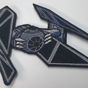 May include: A black and gray embroidered patch of a TIE fighter spaceship from Star Wars. The spaceship is in a flying position with the wings spread out.