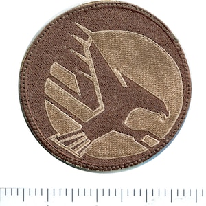 May include: A brown circular patch with a tan embroidered eagle design. The eagle is facing right and has a wing spread. The patch has a stitched border.