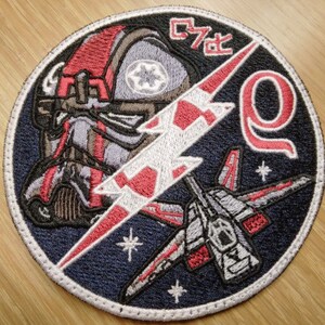 May include: A round, embroidered patch with a blue background. The patch features a red and white lightning bolt, a red and white starfighter, a red and white helmet with a white star, and the text "C7-Q".