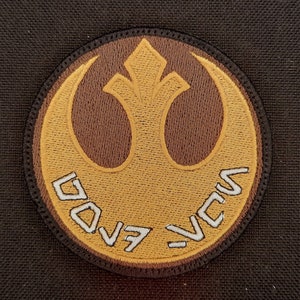 May include: Embroidered patch featuring a circular design with a gold emblem and brown accents. The patch has the text "Rebel" in a stylized font. The patch is likely for a jacket or bag.