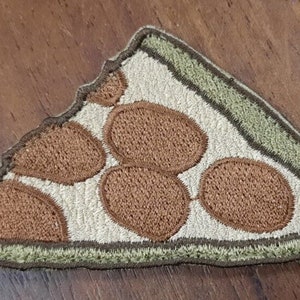 Puede incluir: Un parche bordado con forma de porción de pizza con pepperoni marrón, en colores marrón y verde.