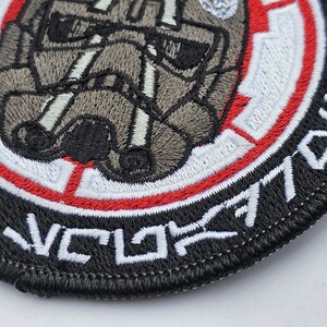 May include: A black and white embroidered patch with a red and white circle in the center. The circle features a gray and white embroidered image of a helmet. The text "V.C.L. 7701" is embroidered around the outside of the circle.