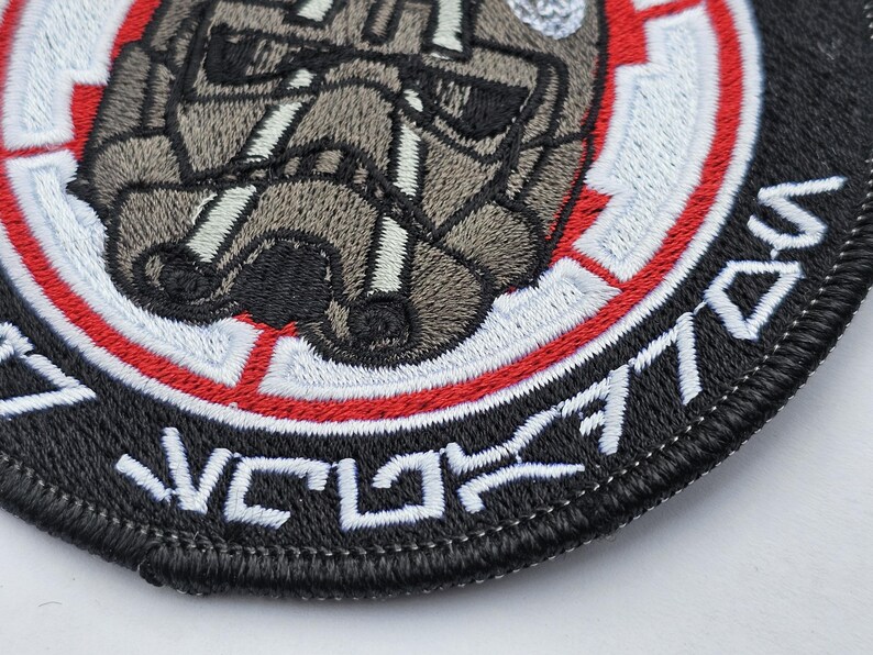May include: A black and white embroidered patch with a red and white circle in the center. The circle features a gray and white embroidered image of a helmet. The text "V.C.L. 7701" is embroidered around the outside of the circle.