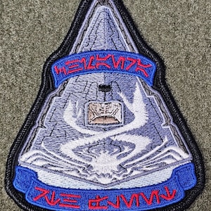 Puede incluir: Un parche bordado gris y blanco con forma de nave espacial con una pancarta azul y roja que dice "HE12K17X" y "71E 82VMV".