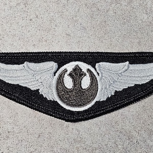 Könnte beinhalten: Gestickter Aufnäher mit schwarzem Rand, einem zentralen Emblem in einem weißen Kreis und silbernen Flügeln. Das Emblem ist dunkelbraun. Dieser Aufnäher ist wahrscheinlich für ein Kostüm oder Kleidungsstück bestimmt. Der Aufnäher ist etwa 10 cm breit.