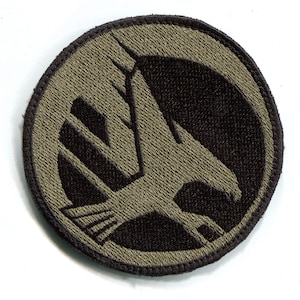 May include: A green and black embroidered patch with a stylized eagle design. The eagle is facing right and has its wings spread. The patch is circular and has a black border.