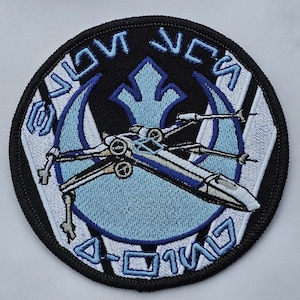 Może przedstawiać: Naszywka haftowana w kolorze czarnym i białym z niebieskim i białym myśliwcem X-wing w środku. Naszywka ma napis "SNOW SPEEDER" i "0-01W17" wokół krawędzi.