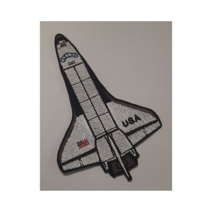 May include: White and black embroidered patch of a space shuttle with the word "USA" and an American flag on the side.