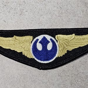 Könnte beinhalten: Gestickter Aufnäher mit schwarzem Rand, goldenen Flügeln und einem blau-weißen Emblem. Der Aufnäher hat ein detailliertes Design und eignet sich für eine Jacke oder Tasche, das ein Symbol der Rebellion darstellt.