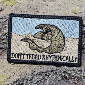 May include: Embroidered patch featuring a brown and tan armadillo-like creature on a beige background with a blue sky. The text "Don't Tread Rhythmically" is embroidered in black at the bottom.