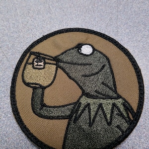May include: A round, brown and black embroidered patch featuring a green frog drinking from a cup. The frog has a white eye and a black mouth. The patch is surrounded by a black border.