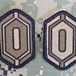 May include: Two olive green and black embroidered patches with a white outline in the shape of a hexagon. The patches have a white number "0" in the center.