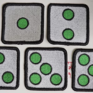 Puede incluir: Seis dados de tela blanca con bordes negros y puntos bordados verdes. Los dados están dispuestos en dos filas de tres.