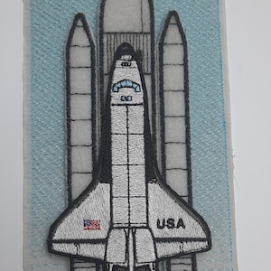 May include: A white and gray embroidered patch of a space shuttle on a blue background. The shuttle has the word "USA" on the side and an American flag on the bottom.