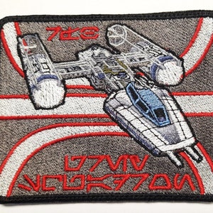 Puede incluir: Un parche gris bordado con una nave espacial blanca y azul en el centro. La nave espacial está rodeada de líneas rojas y blancas. El texto "777V" y "VCLOR770U" está bordado en negro en la parte inferior del parche.