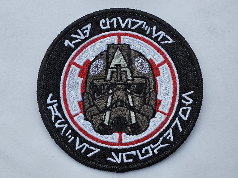 May include: A black and white embroidered patch with a red border. The patch features a stylized helmet with a white symbol and the text "117th  IV  V77  VCL77  IV" in Aurebesh.