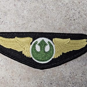 May include: Embroidered patch featuring a black border with gold wings and a green and white emblem. The patch has a unique shape and is likely for a costume or collectible.
