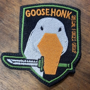 May include: A black and green embroidered patch with a white goose holding a silver knife. The patch says "Goose Honk" and "Special Forces Group".