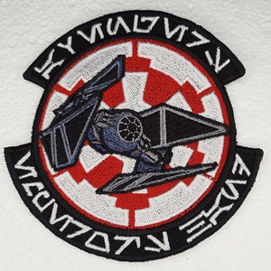May include: Black and white embroidered patch with a red and white imperial symbol and a TIE fighter in the center. The text around the edge reads "KYUULWUVV  VENTO  ERUV".