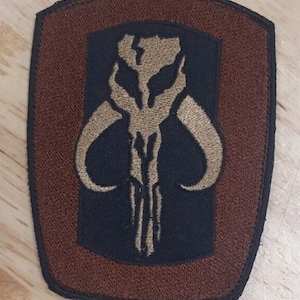 May include: A brown and black embroidered patch with a Mandalorian symbol. The symbol is a stylized skull with horns and a vertical line down the center.