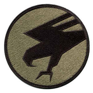 May include: A round, olive green patch with a black embroidered eagle silhouette. The eagle's head is facing left, and its wings are spread out. The eagle's talons are visible.