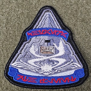 Puede incluir: Un parche bordado gris y blanco con un diseño de nave espacial. El parche tiene una cinta azul con el texto "WE 1CK V7Y" en rojo y otra cinta azul con el texto "77E #1V7V" en rojo.