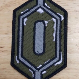 Könnte beinhalten: Ein gestickter Patch in Form eines Sechsecks mit einem großen grauen Buchstaben "O" in der Mitte. Der Patch ist grün, grau und schwarz.