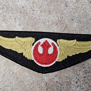 May include: Embroidered patch featuring a gold-colored wing design on a black background. The center of the patch has a red and white emblem. The patch is likely for a costume or collectible.