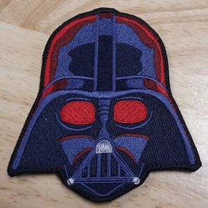 Puede incluir: Un parche bordado de color azul y rojo del casco de Darth Vader. El parche tiene un contorno negro y ojos rojos.