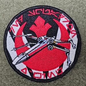 Op de afbeelding: Een geborduurde patch in zwart en wit met een rood Rebel Alliance-symbool en een witte X-wing-jager. De tekst "TWG" en "ROGUE SQUADRON" is geborduurd rond de rand van de patch.