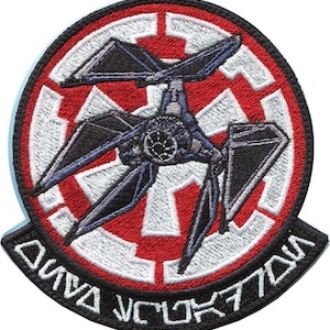 Może przedstawiać: Naszywkę haftowana w kolorze czarnym i białym z czerwonym tłem. Naszywka przedstawia statek kosmiczny TIE Interceptor przed symbolem Imperium. Tekst "DIVA SQUADRON" jest napisany wokół krawędzi naszywki.