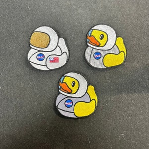 May include: Three embroidered patches featuring yellow duck astronauts in white and gray space suits. Each patch has the NASA logo and a different design. One duck has a brown helmet, and the others have clear visors.