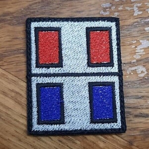 Puede incluir: Un parche blanco bordado con un borde negro. El parche tiene cuatro cuadrados, dos rojos y dos azules. Los cuadrados rojos están en la parte superior y los cuadrados azules en la parte inferior.