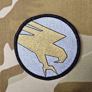 May include: A white and gold embroidered patch with a stylized bird design. The patch is circular and has a black border.