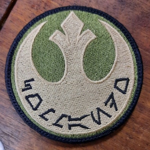 May include: A green and tan embroidered patch featuring the Rebel Alliance symbol and the text "Rebel Alliance" in Aurebesh.