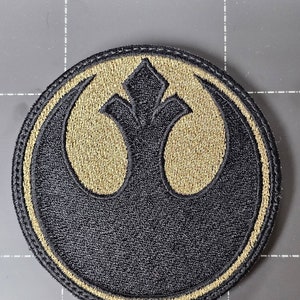 Op de afbeelding: Geborduurd embleem met het symbool van de Rebel Alliance uit Star Wars. Het ronde embleem heeft een zwarte achtergrond met een goudkleurig embleem en een gouden rand. Het embleem is waarschijnlijk van stof.
