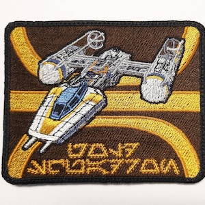 May include: A brown and gold embroidered patch featuring a white and blue spaceship with the text "The Mandalorian" in gold.