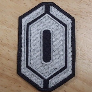 May include: A black and silver embroidered patch shaped like a hexagon with a black letter "O" in the center.