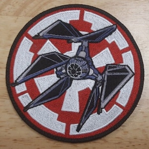 May include: A round embroidered patch featuring a TIE Silencer starfighter in black, blue, and grey, flying over a white Imperial symbol on a red background.