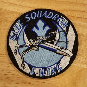 May include: A round, black and white embroidered patch with a blue X-Wing fighter in the center. The patch says "Blue Squadron X-Wing" around the edge.