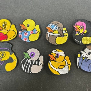Könnte beinhalten: Satz von neun gestickten Bügelbildern mit verschiedenen Designs von gelben Gummienenten. Die Enten tragen verschiedene Kostüme und Accessoires, darunter einen Smoking, einen Pilotenhelm, eine Schiedsrichteruniform und einen Verband.