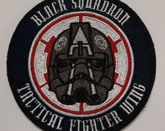 Puede incluir: Un parche bordado en blanco y negro con el texto "Black Squadron Tactical Fighter Wing" en un círculo. El centro del círculo presenta una imagen estilizada de un casco de piloto.