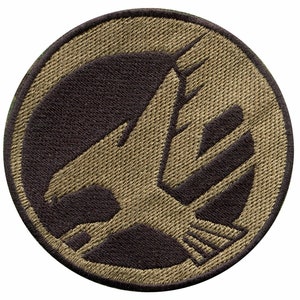 May include: A round, tan-colored patch with a black outline. The patch features a stylized, tan-colored eagle with its wings spread. The eagle is facing left and has a sharp beak.
