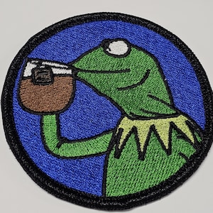Puede incluir: Parche bordado con una rana verde con un ojo blanco, que lleva un collar amarillo, bebiendo de una taza marrón. La rana está sobre un fondo azul con un borde negro.