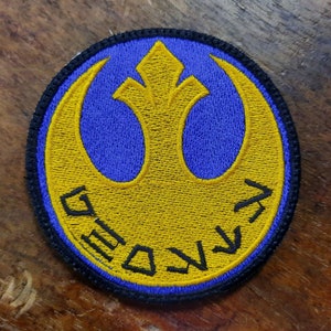 Op de afbeelding: Een gele geborduurde patch met een blauwe achtergrond en een Rebel Alliance-symbool. Het symbool is een gestileerde vogel met uitgespreide vleugels. De tekst "REBEL ALLIANCE" is rond het symbool geborduurd.