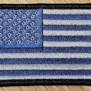 May include: Embroidered patch featuring a stylized American flag design. The patch has a blue field with white stars and blue and white stripes. The border is black.