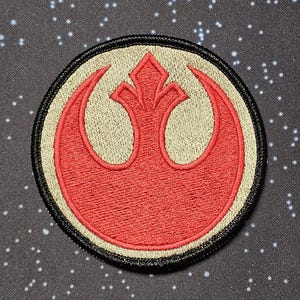 May include: Embroidered patch of the Rebel Alliance symbol from Star Wars. The patch features a red emblem on a beige background, with a black border. The background is a dark grey fabric with white star-like specks.