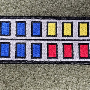 May include: A white and gray embroidered patch with a grid of 12 squares. The squares are filled with blue, yellow, and red.