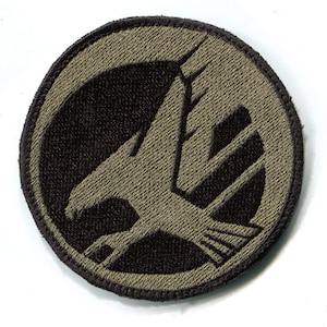 May include: A round, green and brown embroidered patch with a stylized eagle design.
