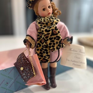 May include: A Madame Alexander doll dressed in a pink coat, leopard print scarf and hat, and brown boots. The doll holds a brown purse and a shopping bag. A tag reads "Madame Alexander Classic The Fashion Collection Platinum."